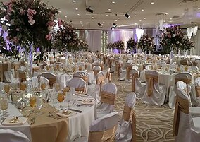 A banquet hall with round tables covered in white tablecloths, adorned with floral centerpieces and white chairs with gold sashes.