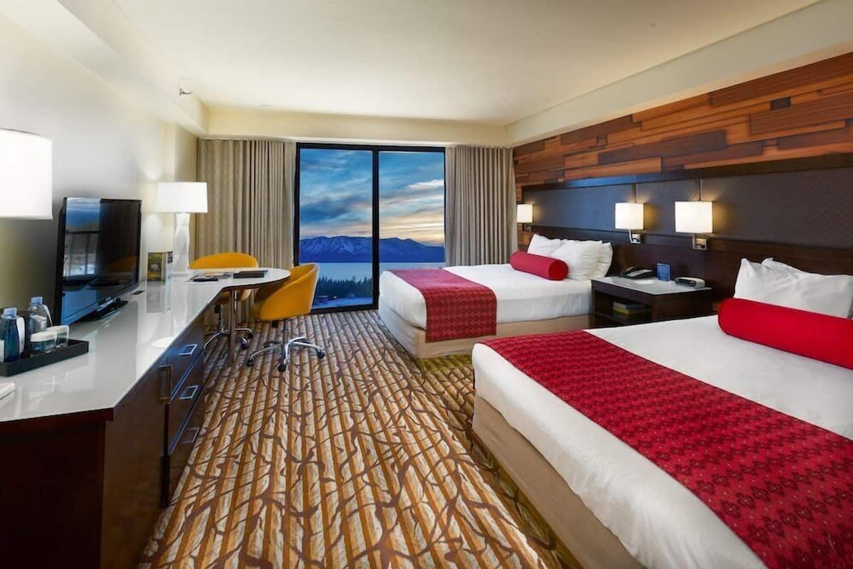 Quarto de hotel com escrivaninha, duas camas, televisão e uma janela grande com vista para as montanhas.