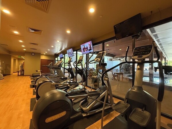 Une rangée de vélos d’appartement dans une salle de sport, avec un plancher en bois et de grandes fenêtres.