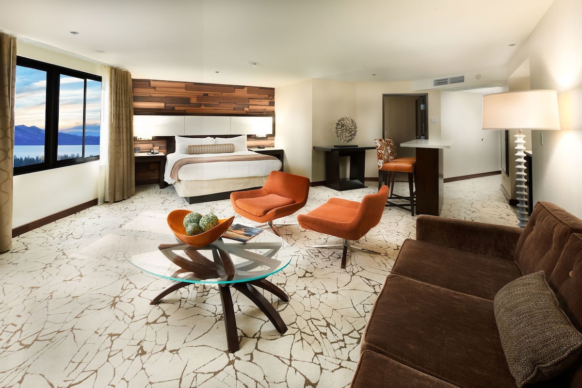Ein Hotelzimmer mit einem Bett, zwei orangefarbenen Sesseln, einem Glastisch, einem braunen Sofa, einer Lampe und Blick auf Berge durch große Fenster.