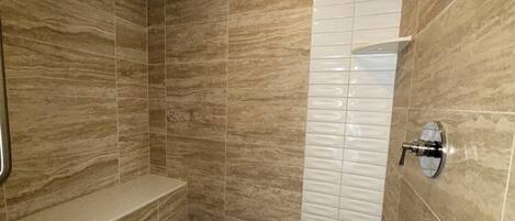 Un baño con paredes de mármol color beige, zona de ducha con azulejos blancos y un banco empotrado.