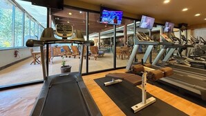 Een sportschool met loopbanden, een bank en trainingsapparatuur. Er zijn grote ramen en een televisiescherm.