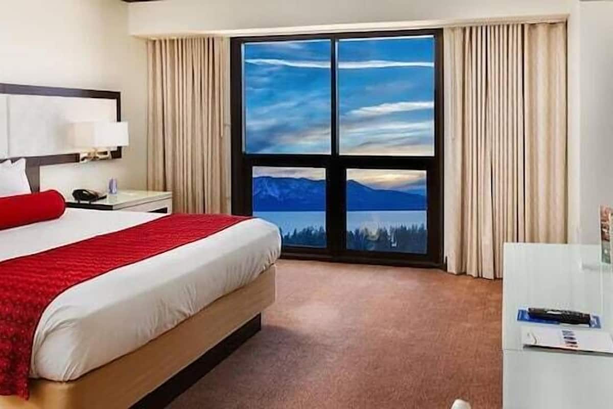 Quarto de hotel com uma cama, uma mesa, uma televisão e uma grande janela com vista para as montanhas.