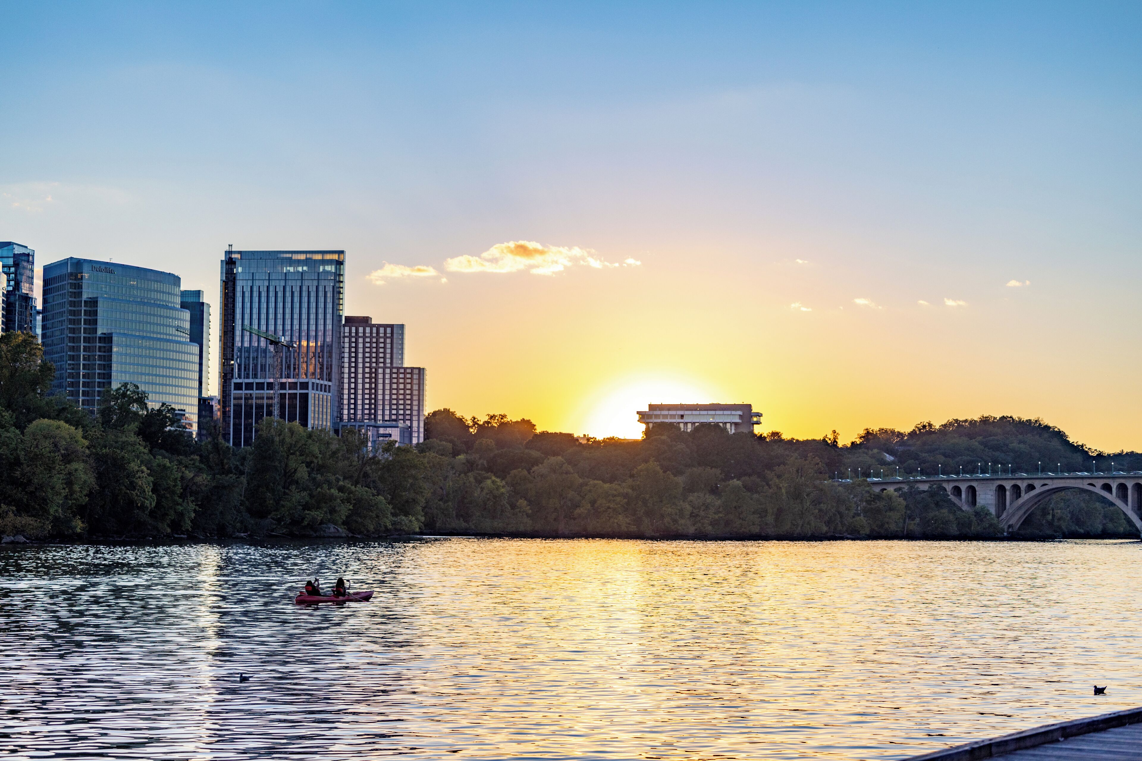A city skyline with modern buildings, a bridge, and a sunset over a body of water.