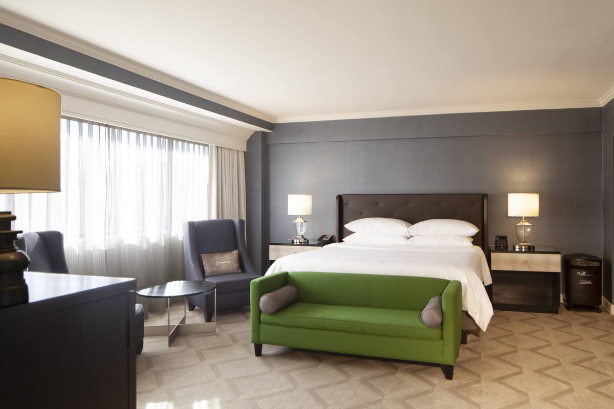 A hotel room with a large bed, two bedside tables with lamps, a green sofa, a chair, and a desk with a lamp.