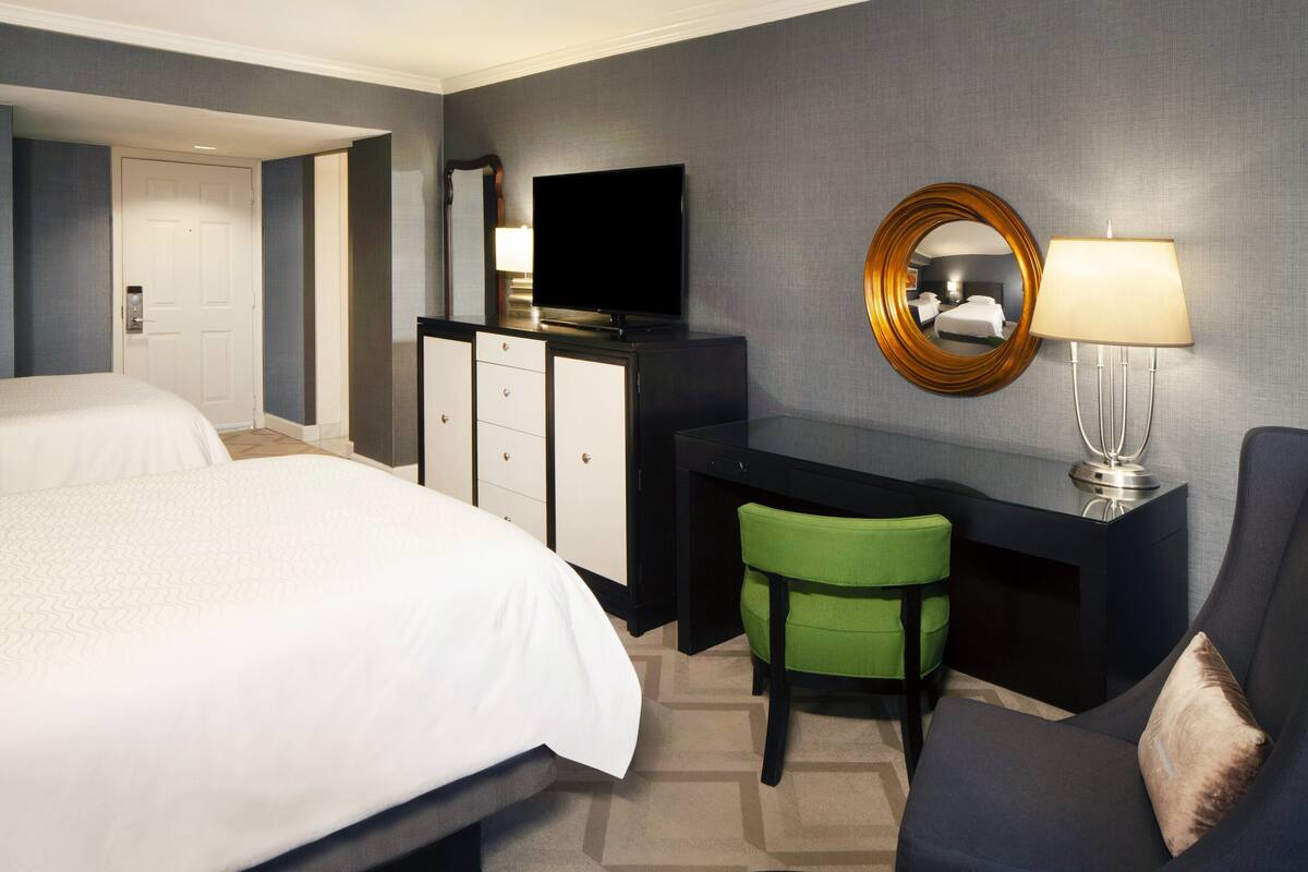A hotel room with two beds, a desk with a television, a mirror, and a lamp.