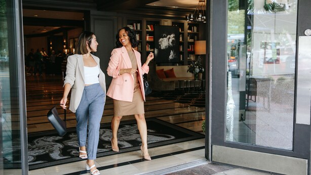 Dos mujeres caminando por el vestíbulo de un hotel con un amplio ventanal, una mesa de recepción y una zona de estar con un sofá y una mesa de centro.