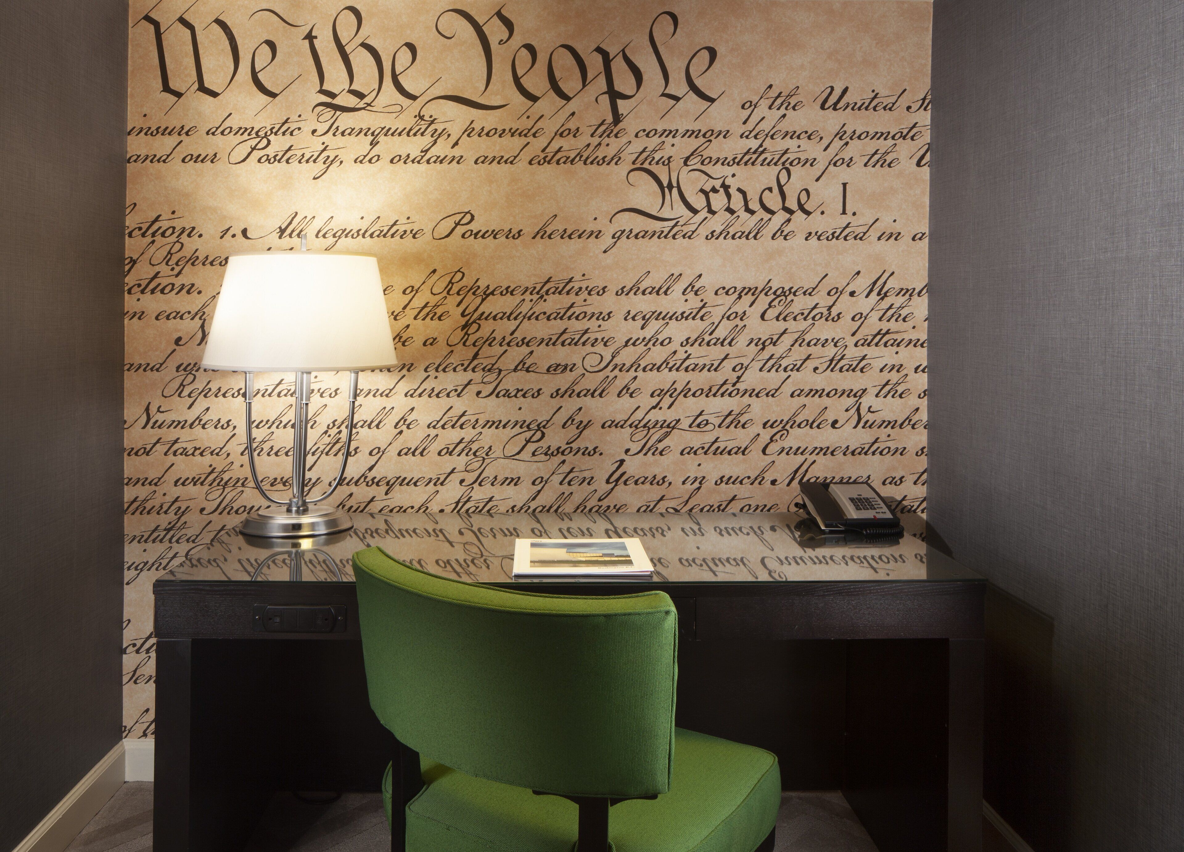 A desk with a green chair, a lamp, and a book. The wall behind features a large, decorative script.