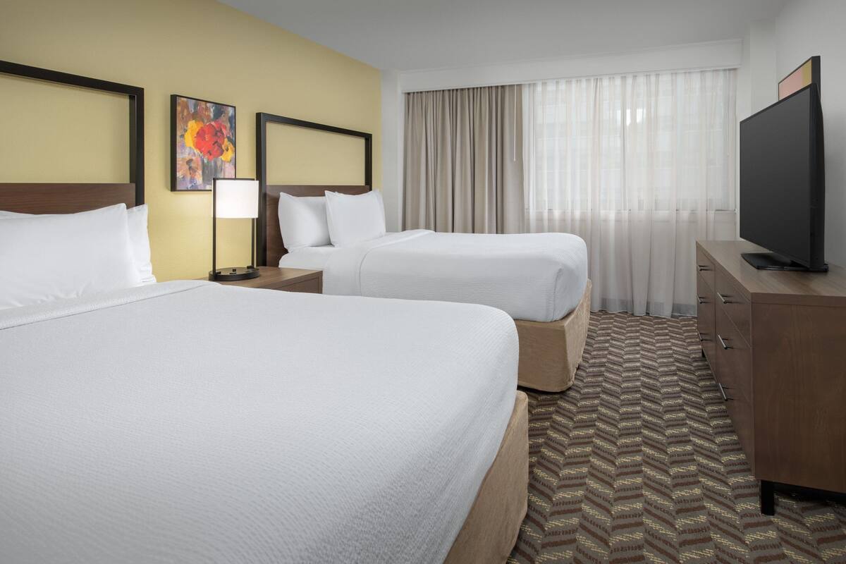 A hotel room with two beds, a television, a desk, and a window with curtains.