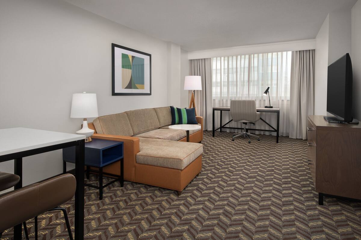 A hotel room with a sofa, a desk, a chair, a television, and a window with curtains.