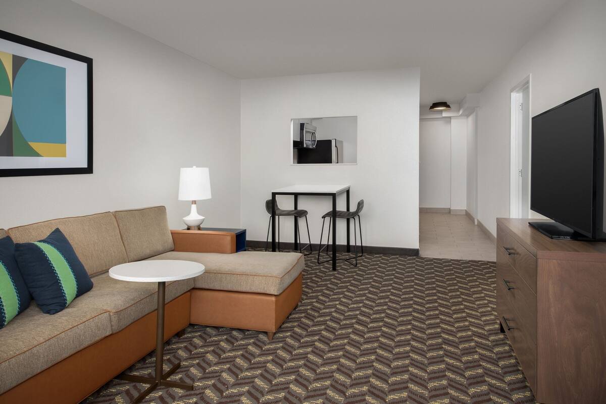 A modern hotel room with a sofa, a small round table, a TV, and a kitchenette area.