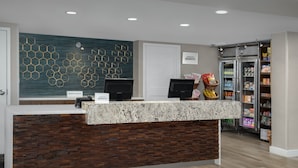 Ein Hotel-Rezeptionsbereich mit modernem Design, darunter ein schlicht gestalteter Empfangstresen, eine dekorative Wand und eine Selbstbedienungsgetränke-Maschine.