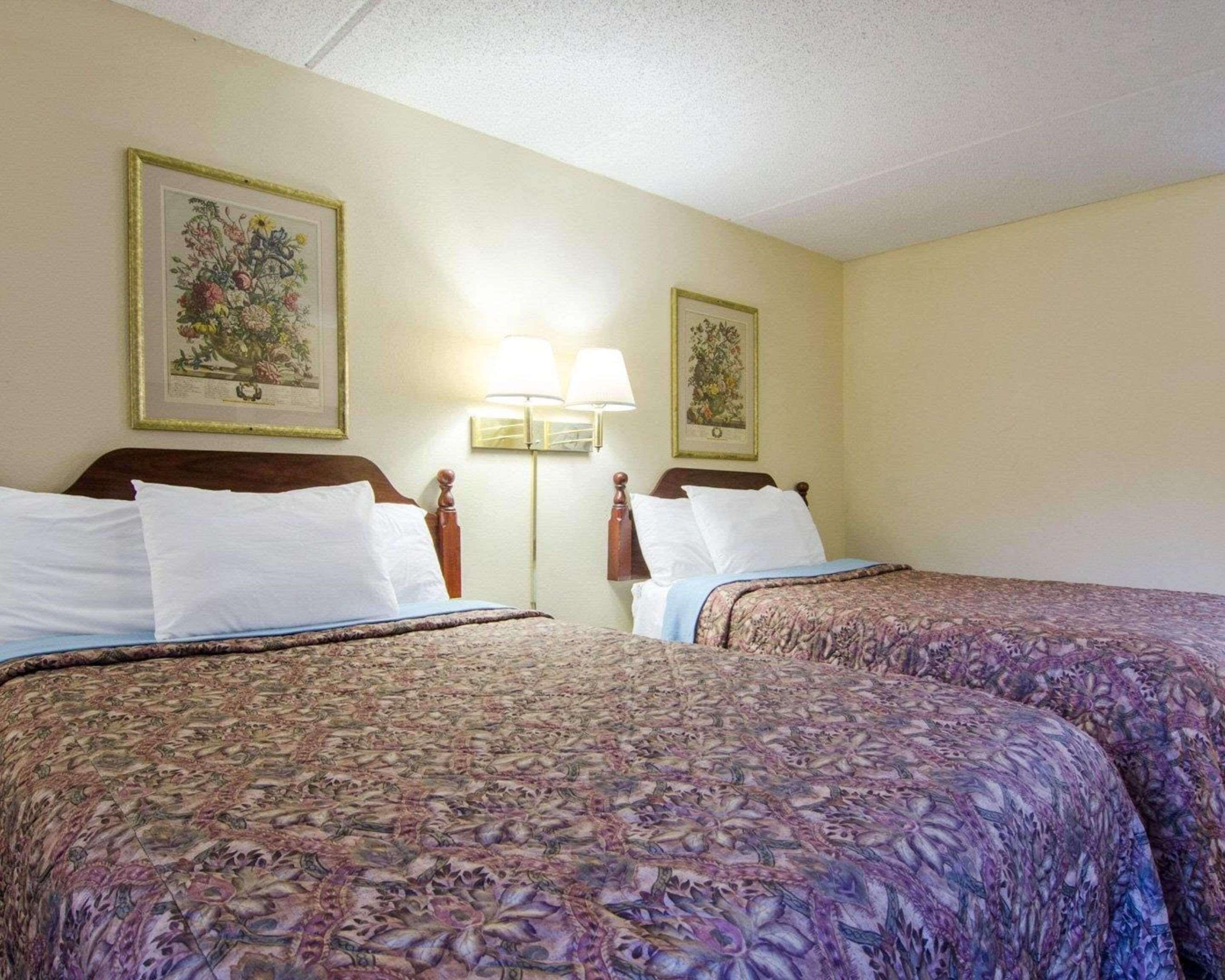 A hotel room with two beds, each with a floral-patterned bedspread, a wooden headboard, and white pillows. There are two wall-mounted lamps between the beds.