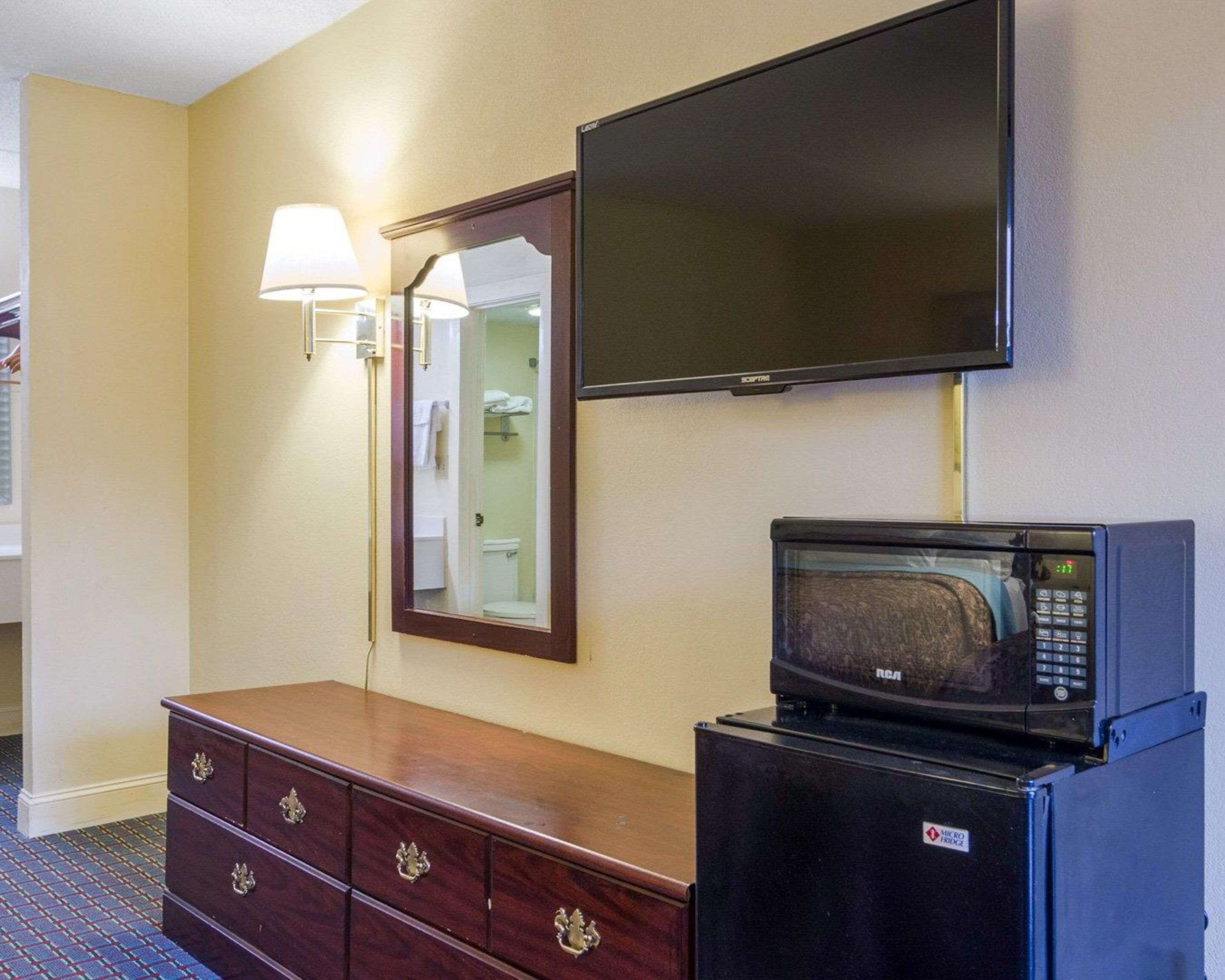A hotel room with a flat-screen TV mounted on the wall, a microwave on a small table, a wooden dresser with drawers, and a mirror with a wall-mounted lamp.