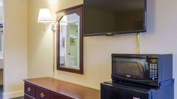 Une chambre d’hôtel équipée d’un téléviseur à écran plat fixé au mur, d’un micro-ondes posé sur une petite table, d’une commode en bois avec des tiroirs et d’un miroir avec une lampe murale.