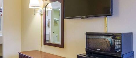 A hotel room with a flat-screen TV mounted on the wall, a microwave on a small table, a wooden dresser with drawers, and a mirror with a wall-mounted lamp.