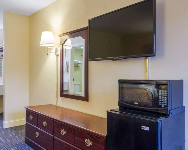 A hotel room with a flat-screen TV mounted on the wall, a microwave on a small table, a wooden dresser with drawers, and a mirror with a wall-mounted lamp.