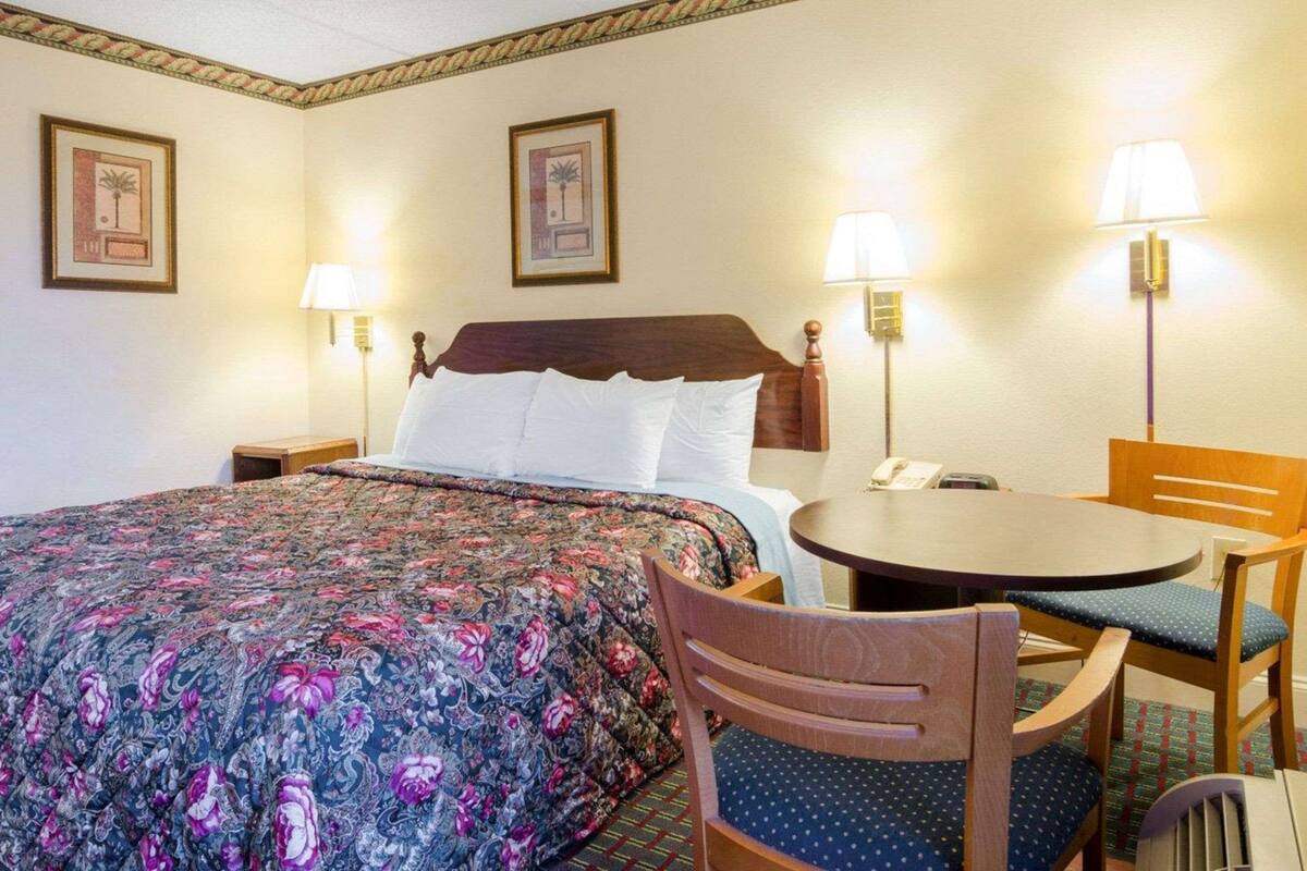 A hotel room with a bed, two chairs, a small table, and two wall-mounted lamps.
