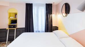 Une chambre d’hôtel avec un lit, un petit bureau avec un ordinateur portable, une étagère remplie de livres et une fenêtre avec des rideaux.
