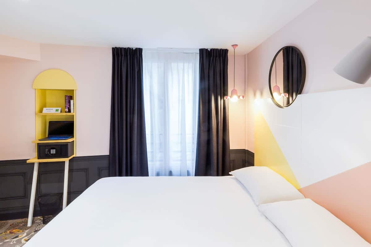 Een hotelkamer met een bed, een klein bureau met een laptop, een plank met boeken en een raam met gordijnen.