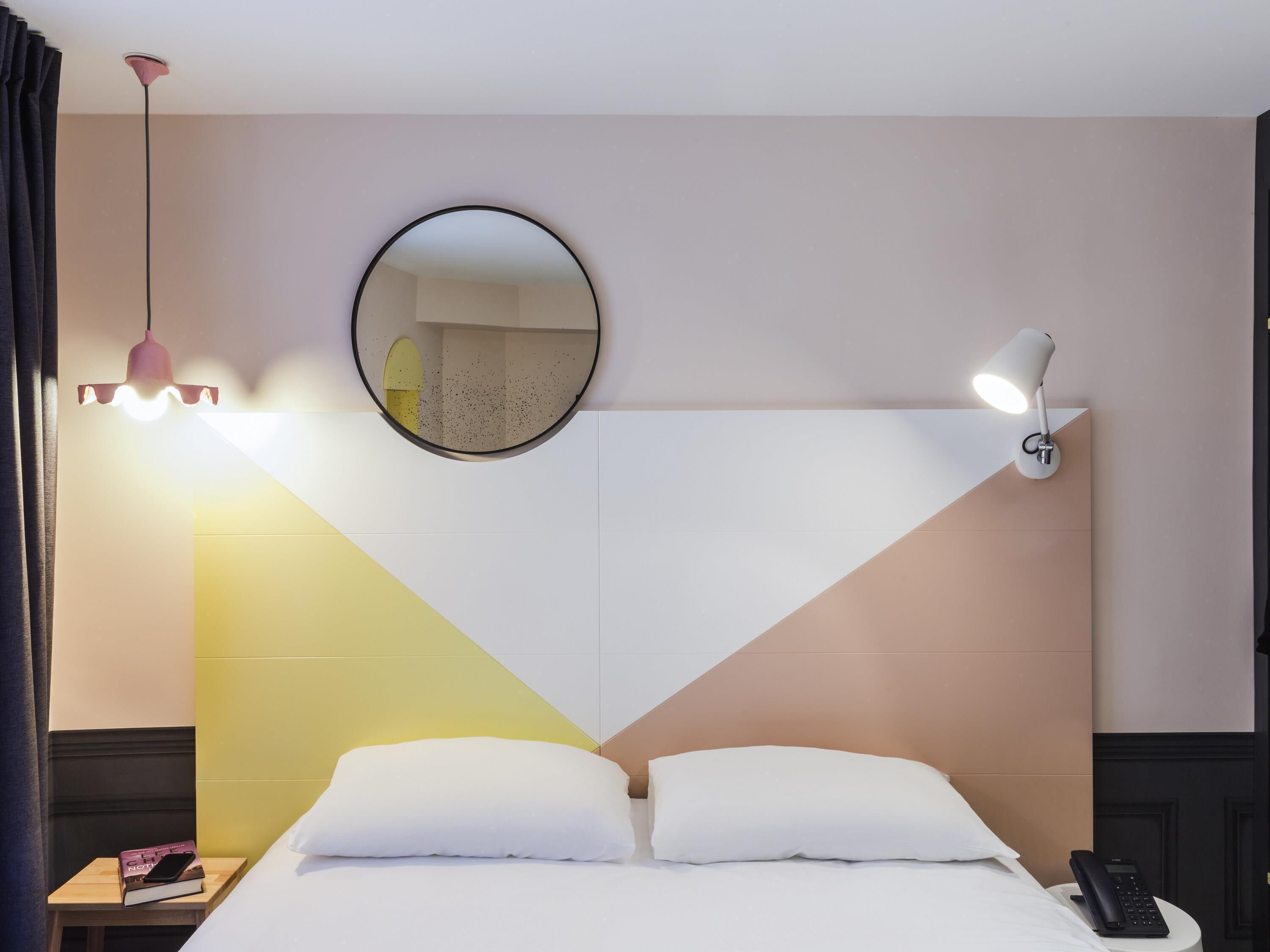 A hotel room with a bed, a bedside table with a phone, a lamp, and a mirror on the wall.