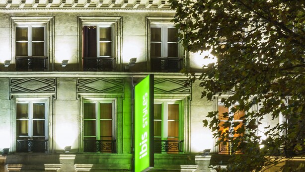 Un bâtiment doté de nombreuses fenêtres et de balcons, illuminé par une enseigne verte sur laquelle on peut lire « ibis styles ».