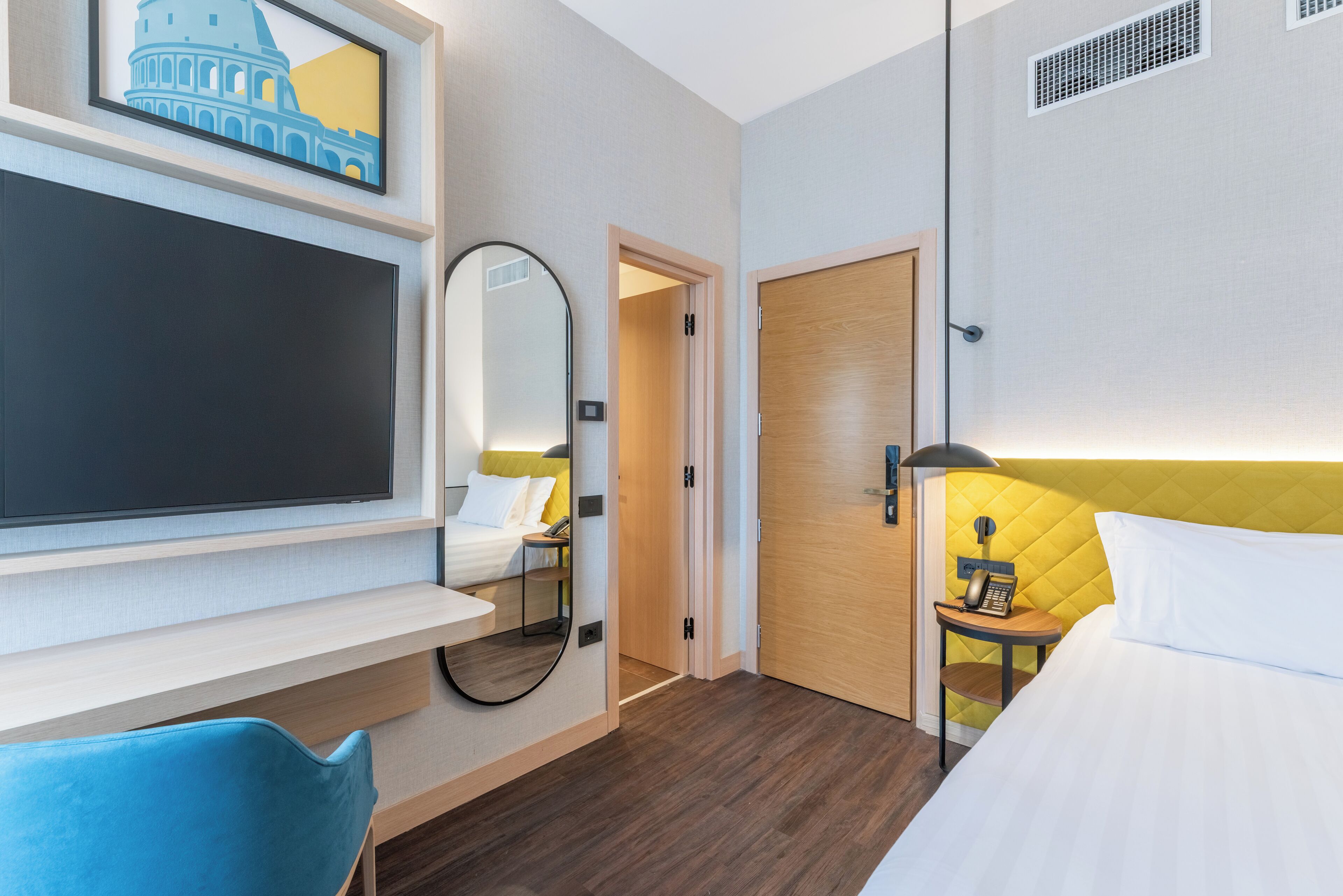Une chambre d’hôtel comprenant un lit, un bureau avec un téléviseur à écran plat, un miroir, une table de chevet avec une lampe et une porte donnant sur une autre pièce.