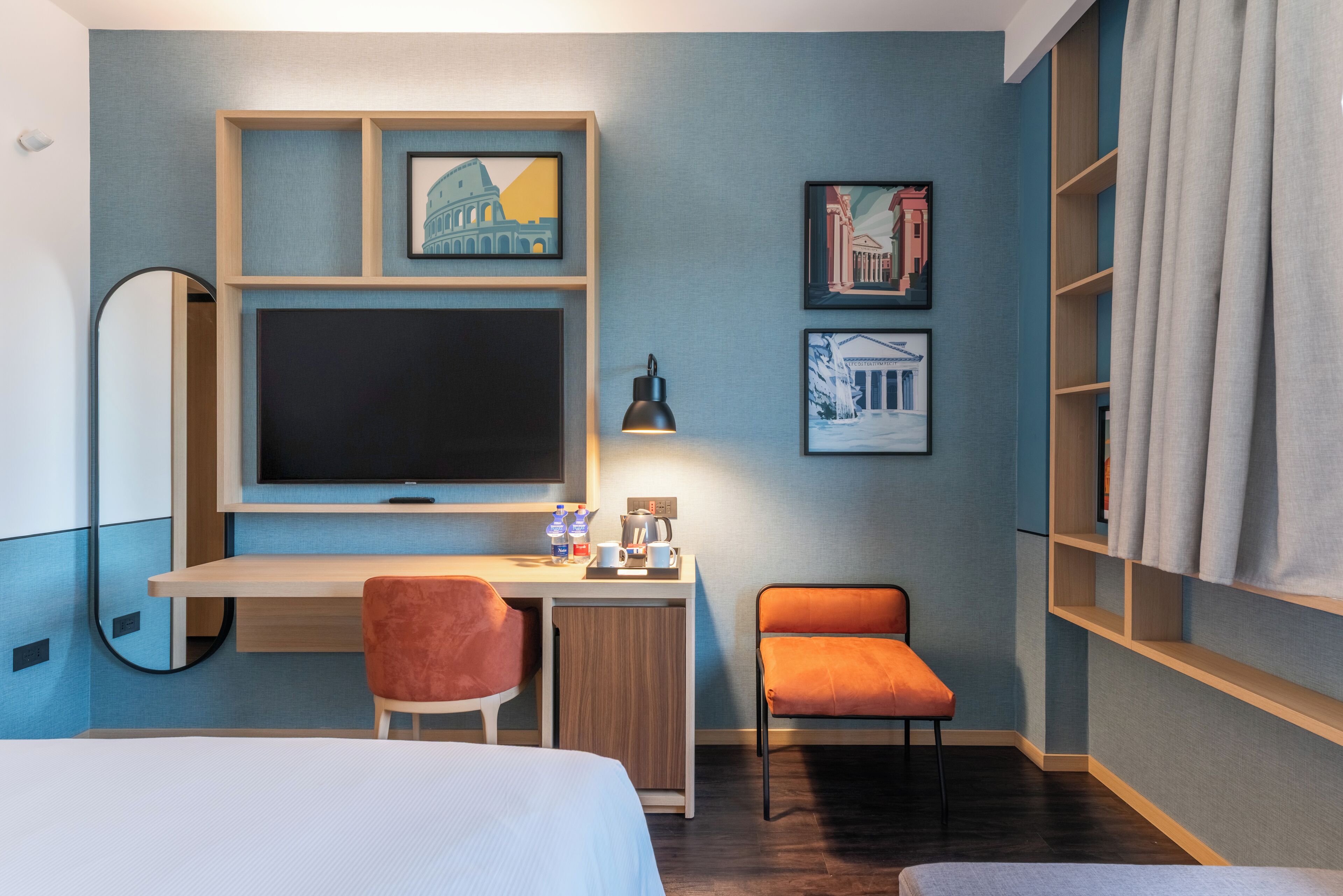 Quarto de hotel com cama, secretária com computador, cadeira, pequena mesa com cafeteira e quadros nas paredes.