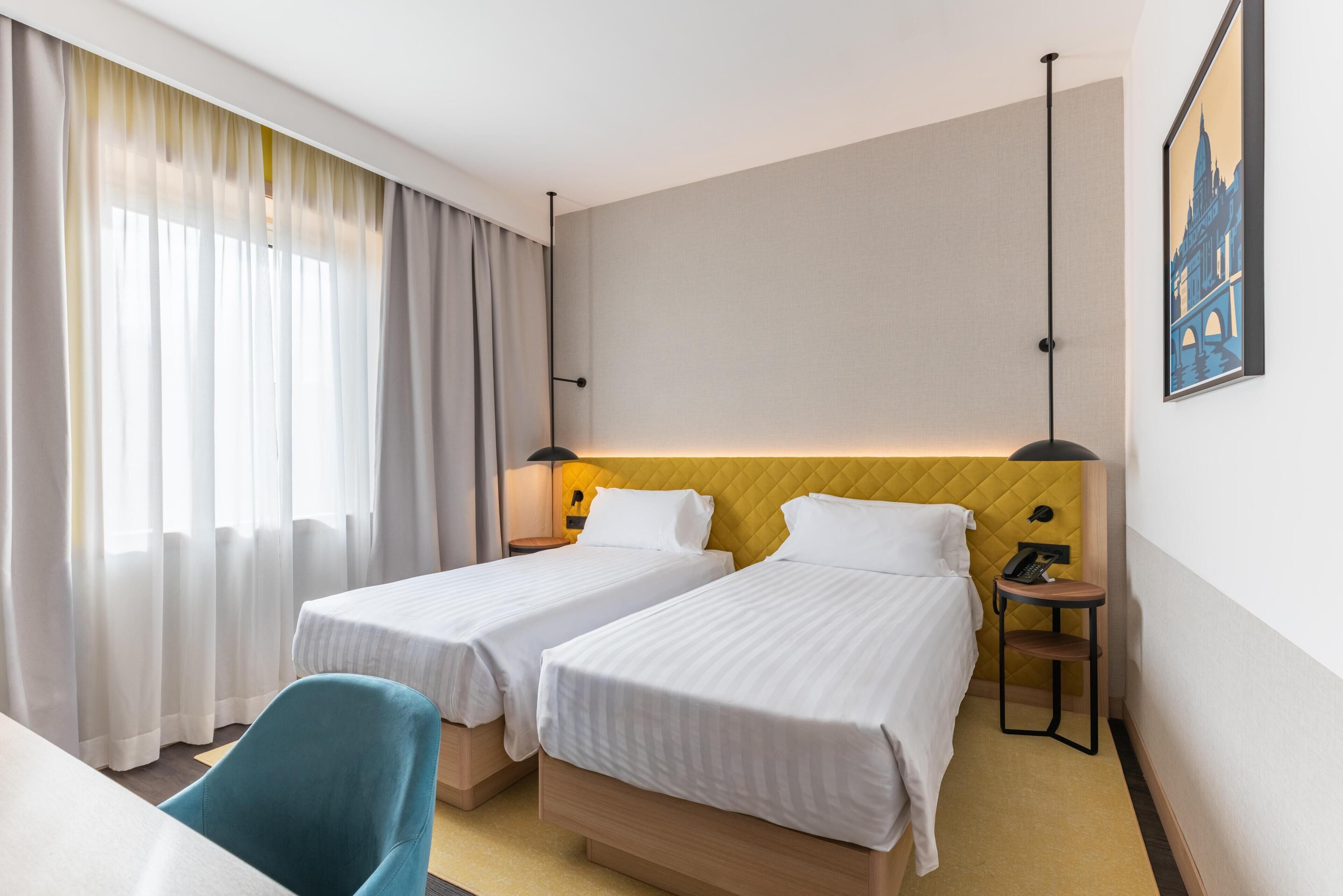 A hotel room with two single beds, a desk, a chair, a nightstand with a phone, and a painting on the wall.