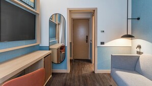 A modern hotel room with a flat-screen TV, a mirror, a sofa, a desk, and a door leading to another room. - Hilton Garden Inn Rome Colosseum (Rome)
