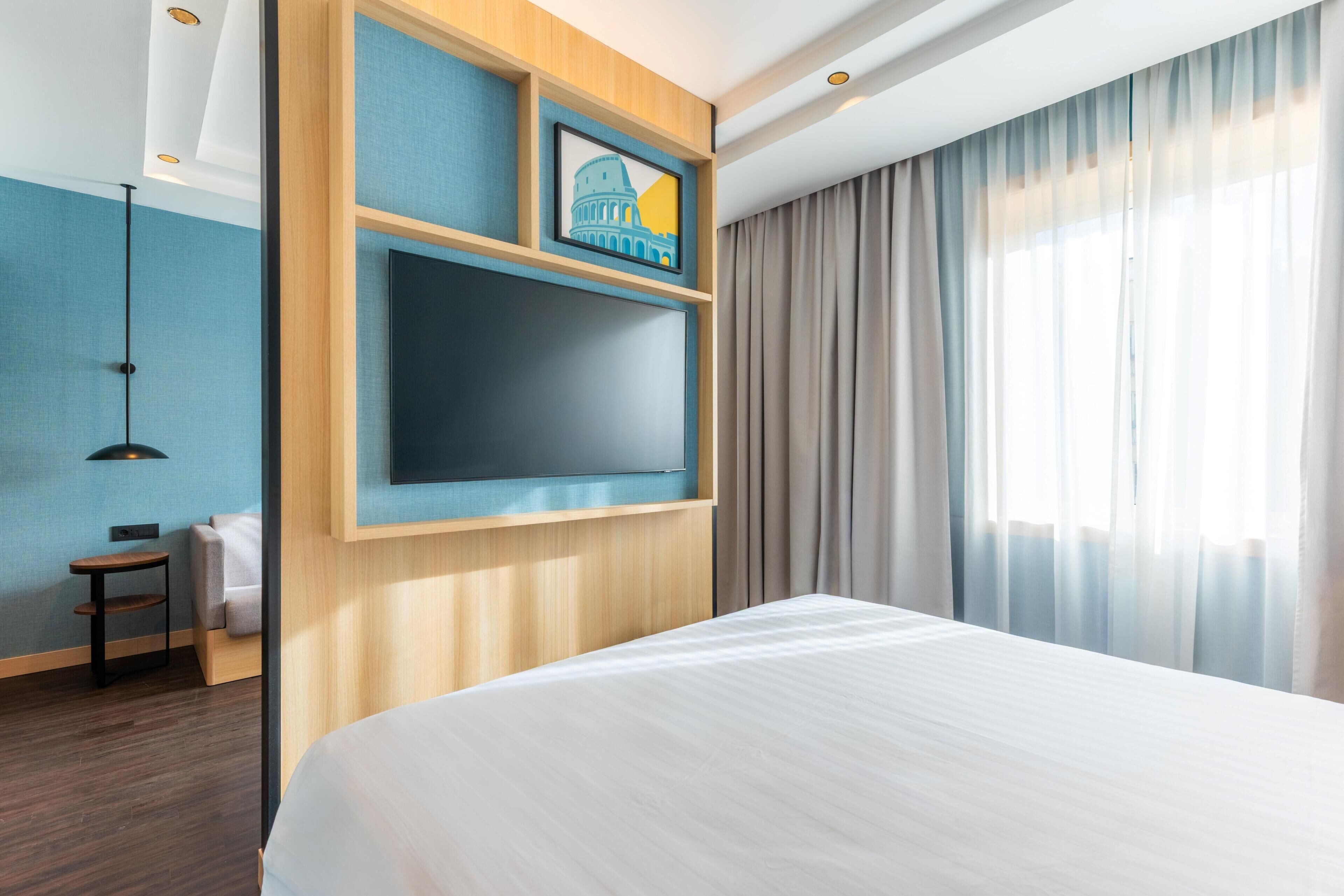Une chambre d’hôtel avec un grand lit, un téléviseur fixé au mur, une petite table et une fenêtre avec des rideaux.