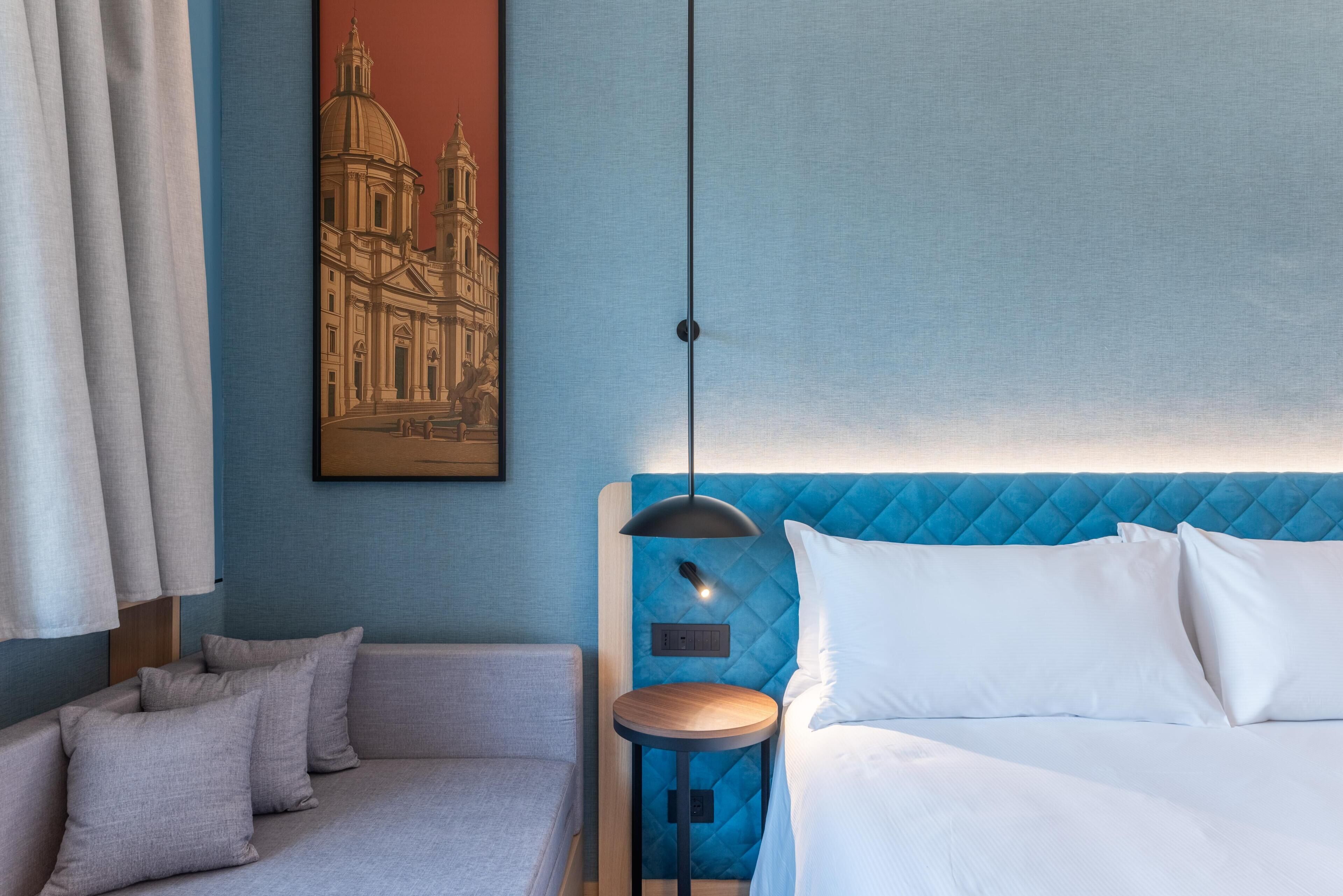 A hotel room with a bed, a small round table, a sofa, and a painting of a building on the wall.