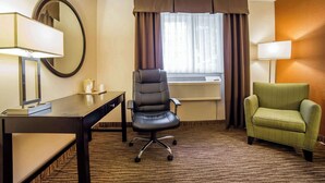 A hotel room with a desk, a green armchair, a black office chair, a round mirror, and a window with curtains.