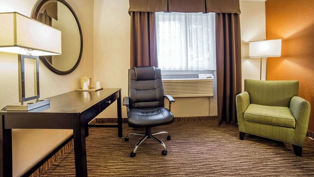 Habitación de hotel con escritorio, un sillón verde, una silla de oficina negra, un espejo redondo y una ventana con cortinas.