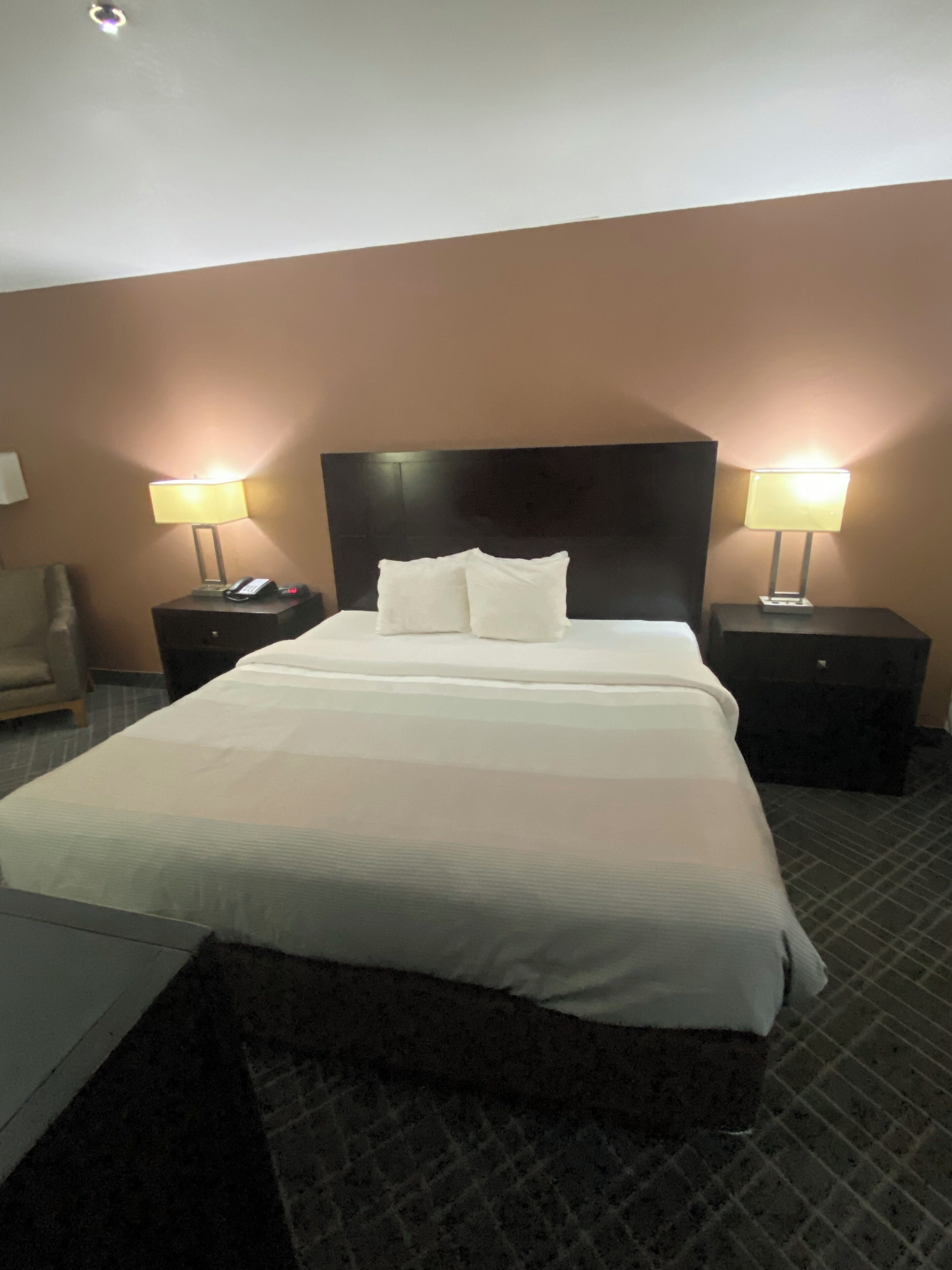 A hotel room with a large bed, two bedside tables with lamps, a chair, and a carpeted floor.