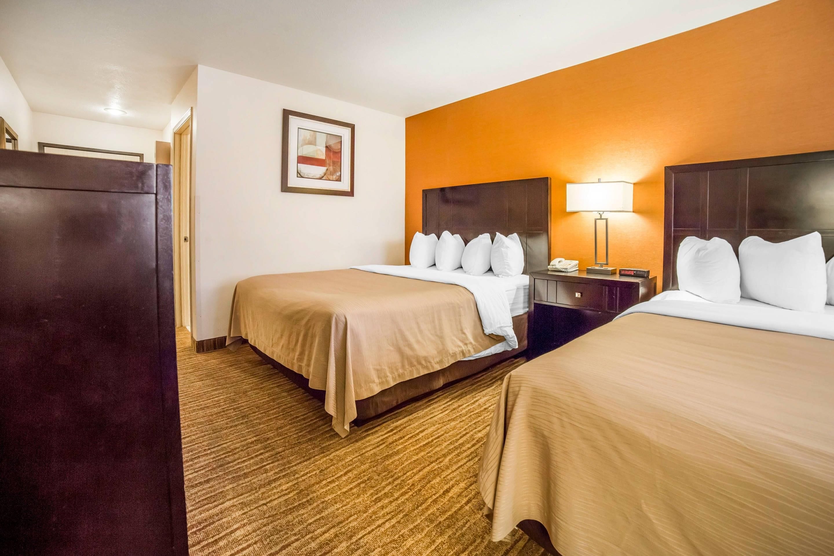 Habitación de hotel con dos camas, una mesita de noche, una lámpara, un cuadro enmarcado en la pared y piso alfombrado.