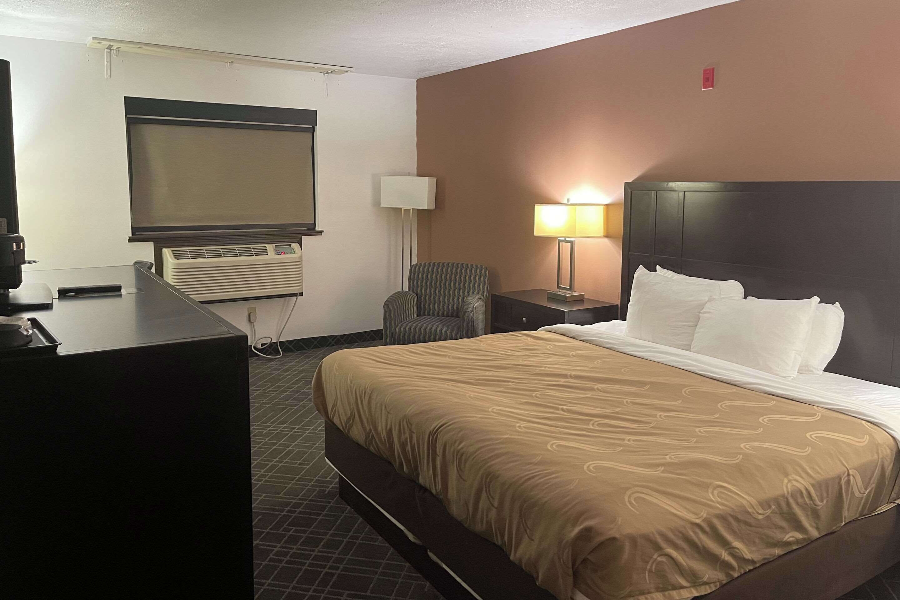 Ein Hotelzimmer mit einem großen Bett, einem Schreibtisch mit Computer, einem Sessel, einer Lampe und einem Fernseher.