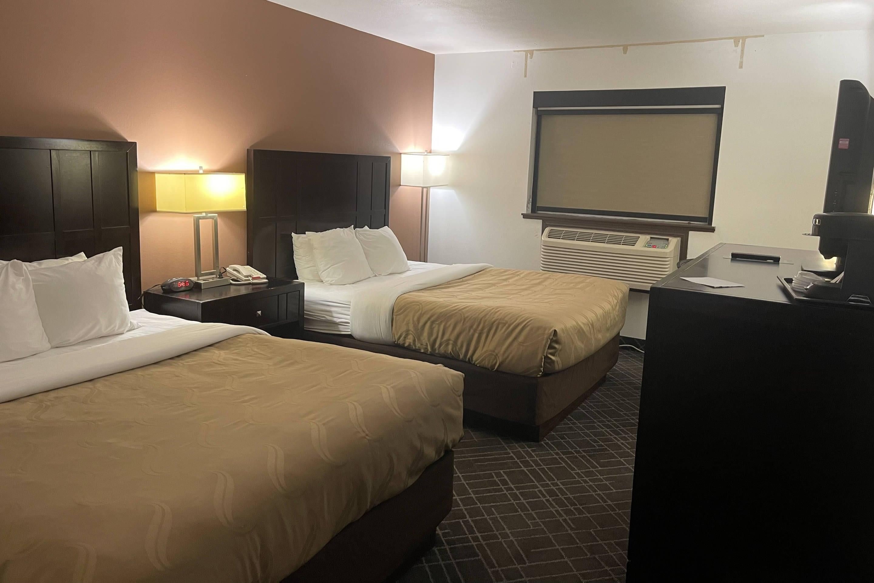 A hotel room with two beds, bedside tables, lamps, a TV, an air conditioning unit, and a desk with a coffee machine.