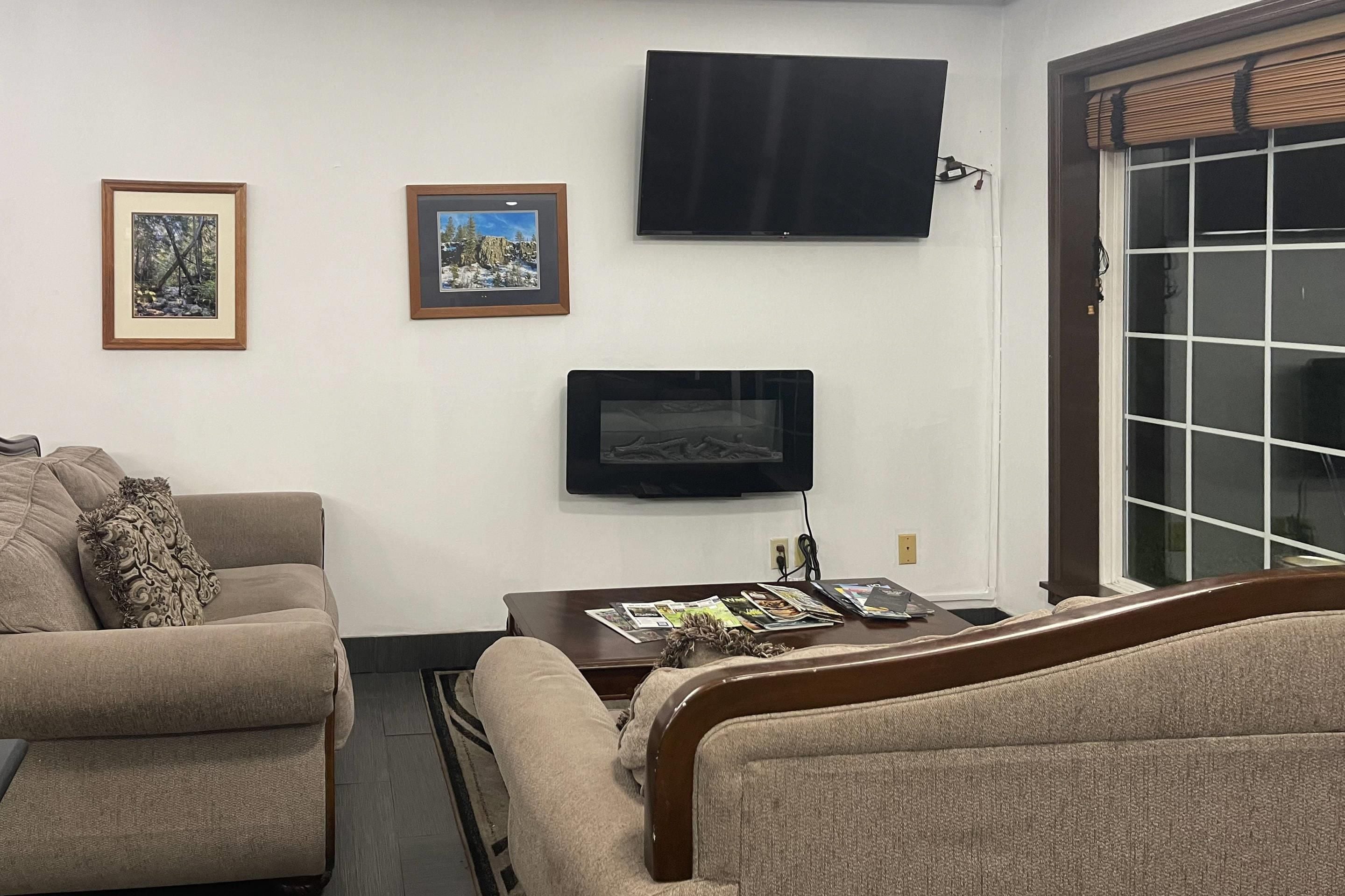 A living room with a sofa, a coffee table with magazines, a flat-screen TV mounted on the wall, and a window with blinds.