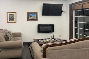 Una sala de estar con sofá, mesa de centro con revistas, televisor de pantalla plana montado en la pared y ventana con persianas.