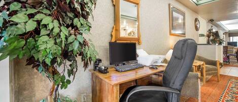 A home office with a wooden desk, a computer, and a chair. There is a large mirror on the wall and a plant on the left.