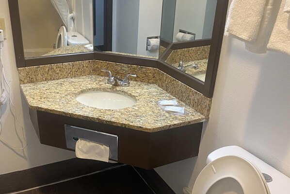 A bathroom with a granite countertop sink, a mirror, a towel dispenser, and a toilet.