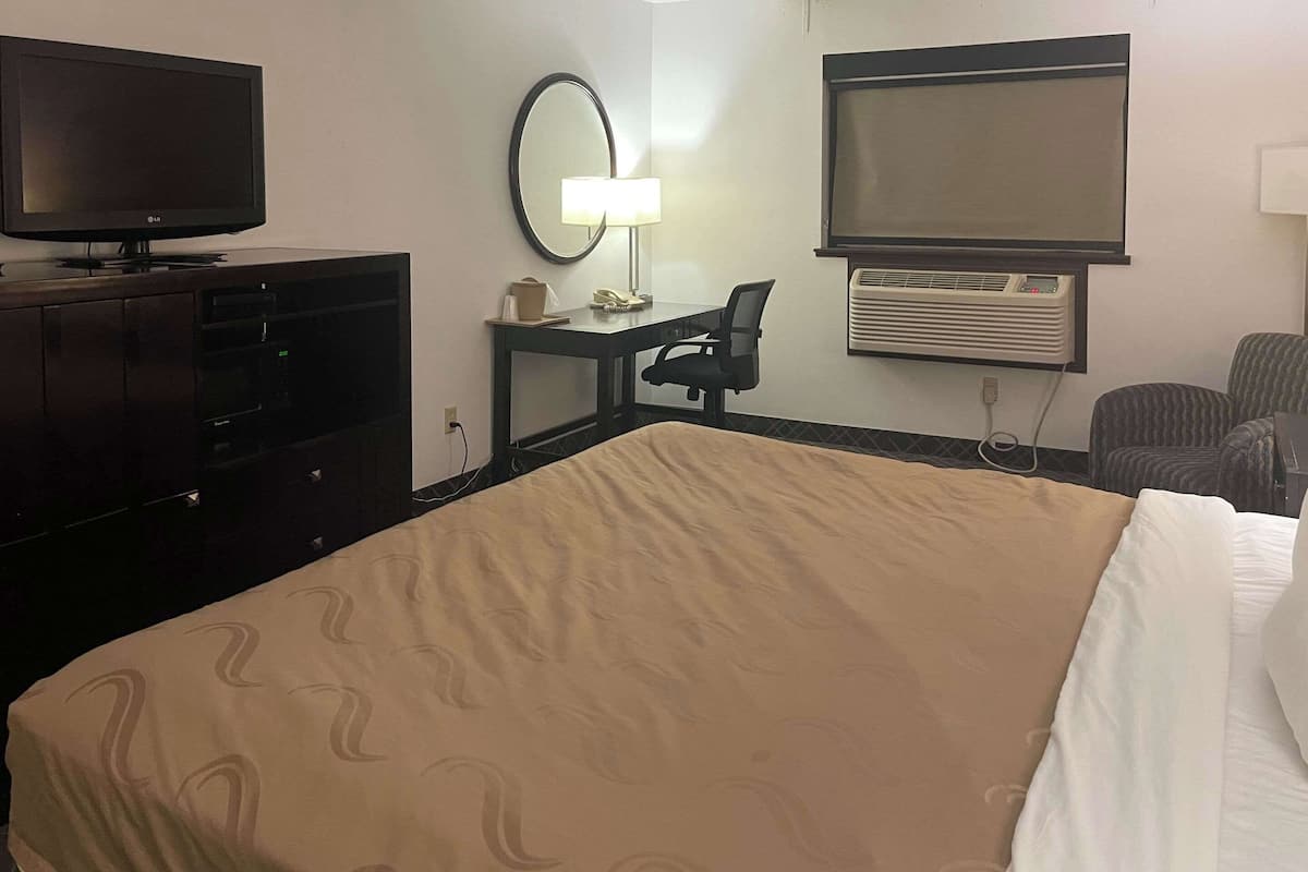 Habitación de hotel con cama, escritorio con silla, televisor, espejo, equipo de aire acondicionado y ventana.