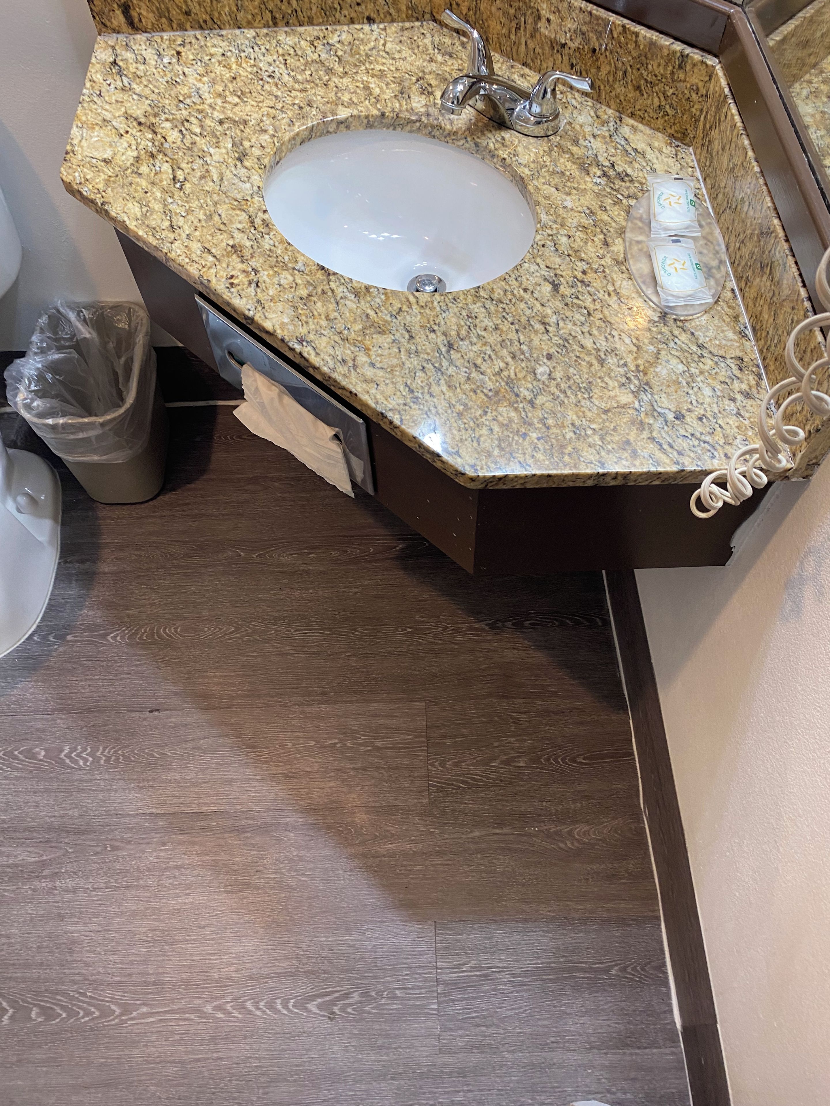 A bathroom with a granite countertop sink, a trash can, and a wooden floor.