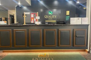 A hotel reception desk with a 'Quality Inn' sign, a reception counter, and a 'Quality' logo on the floor mat.