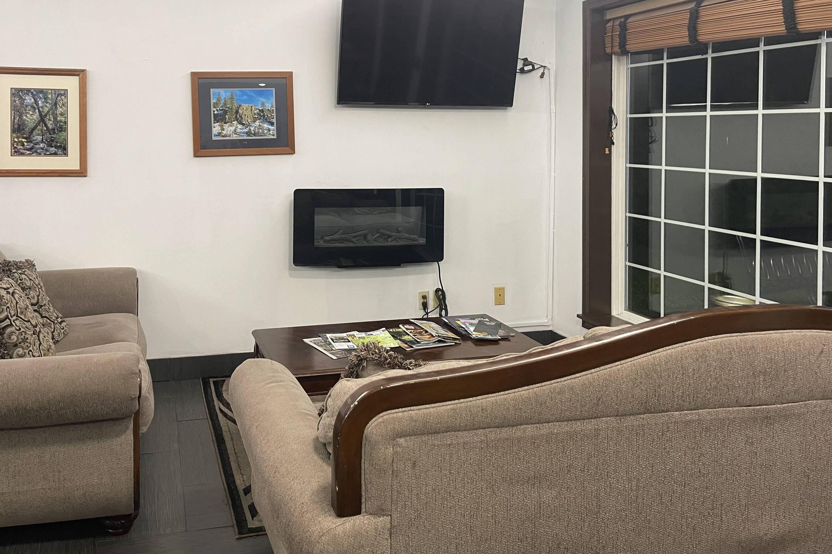 Un salon avec deux canapés, une table basse avec des magazines, un téléviseur à écran plat fixé au mur et une grande fenêtre avec des stores.