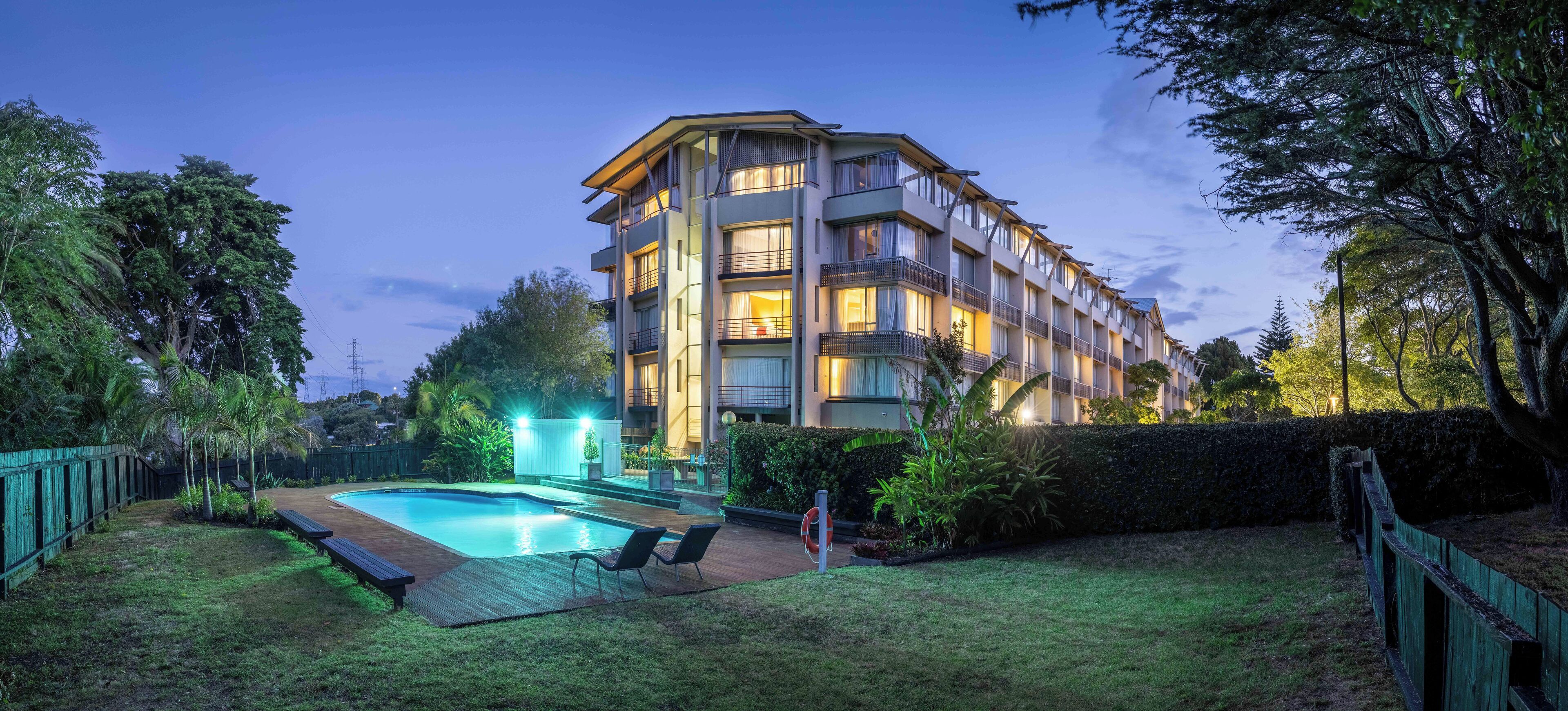 Un hotel de varios pisos con piscina, rodeado de follaje y una terraza de madera.