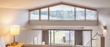 A hotel room with a bed, a nightstand with a lamp, a window with blinds, and a view of the outside.