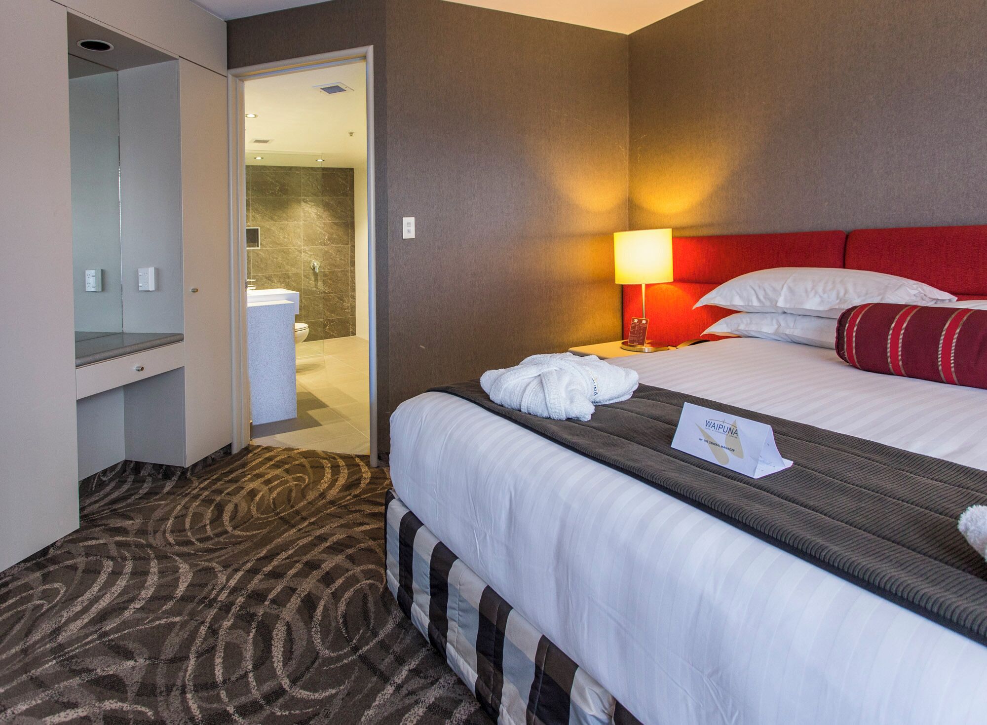 A hotel room with a bed, a nightstand with a lamp, a robe, and a bathroom visible through an open door.