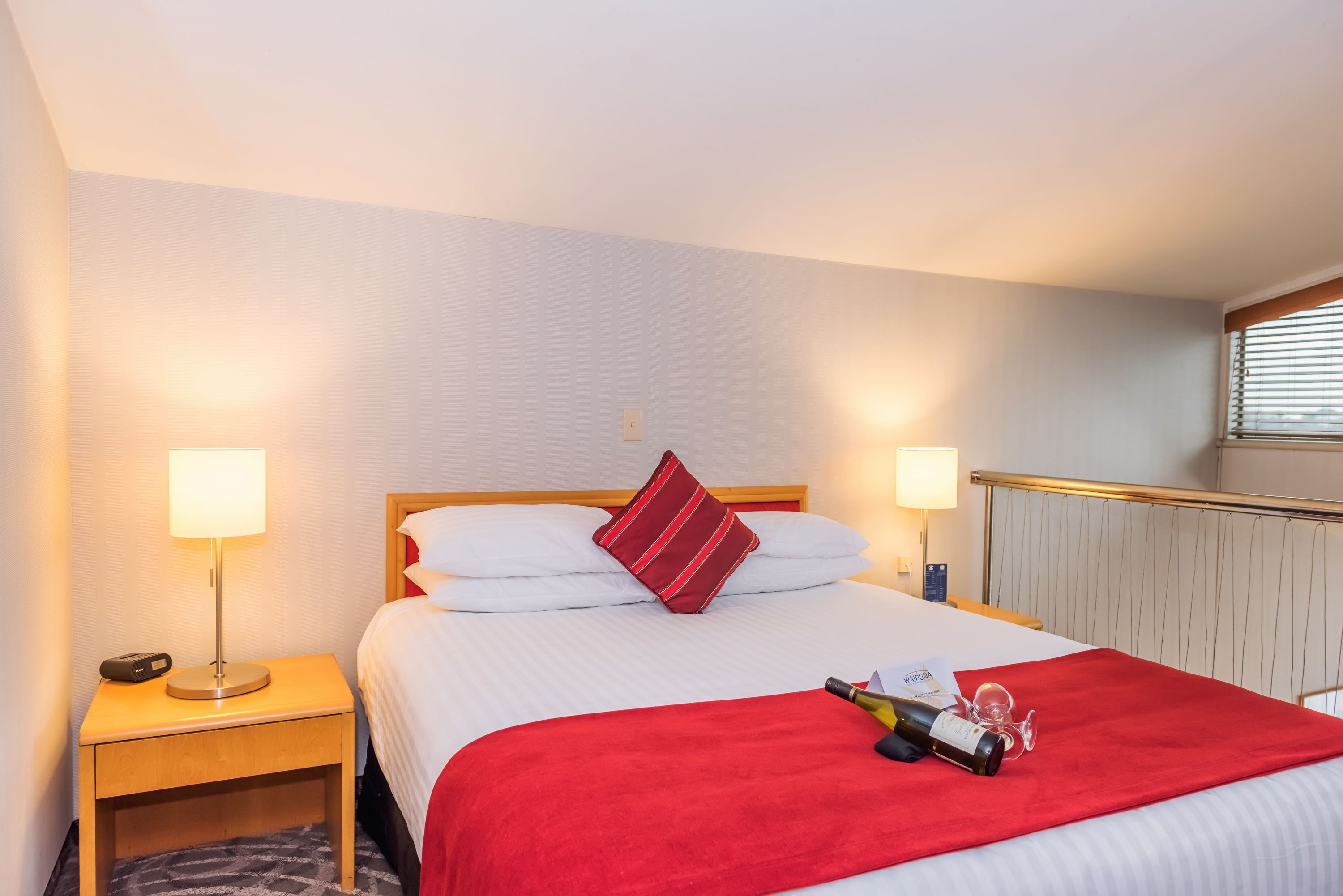 Hotelkamer met een net opgemaakt bed, een rode plaid, een fles champagne en een glas op het bed. Twee bedlampen, een houten nachtkastje en een raam met jaloezieën zijn ook aanwezig.
